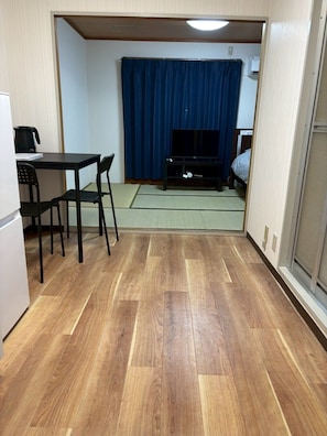 Une pièce avec un plancher en bois, une petite table entourée de chaises, un téléviseur et un rideau bleu.