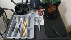 Ein Tisch mit einer schwarzen Teekanne, einer schwarzen Pfanne, einer Schere, einem Messer, Essstäbchen, einem Löffel und einer Tablett mit Besteck.