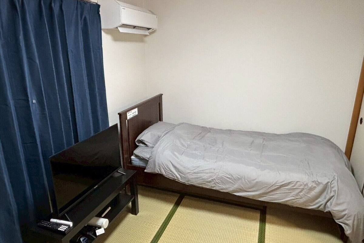 Una habitación pequeña con una cama, un televisor sobre un mueble bajo, un control remoto y un aire acondicionado.