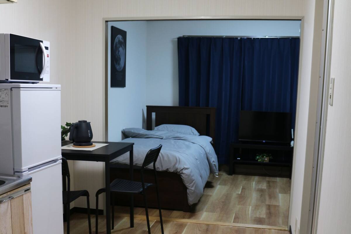 Una habitación compacta con kitchenette, una cama con cortina azul, televisión y una alfombra pequeña.