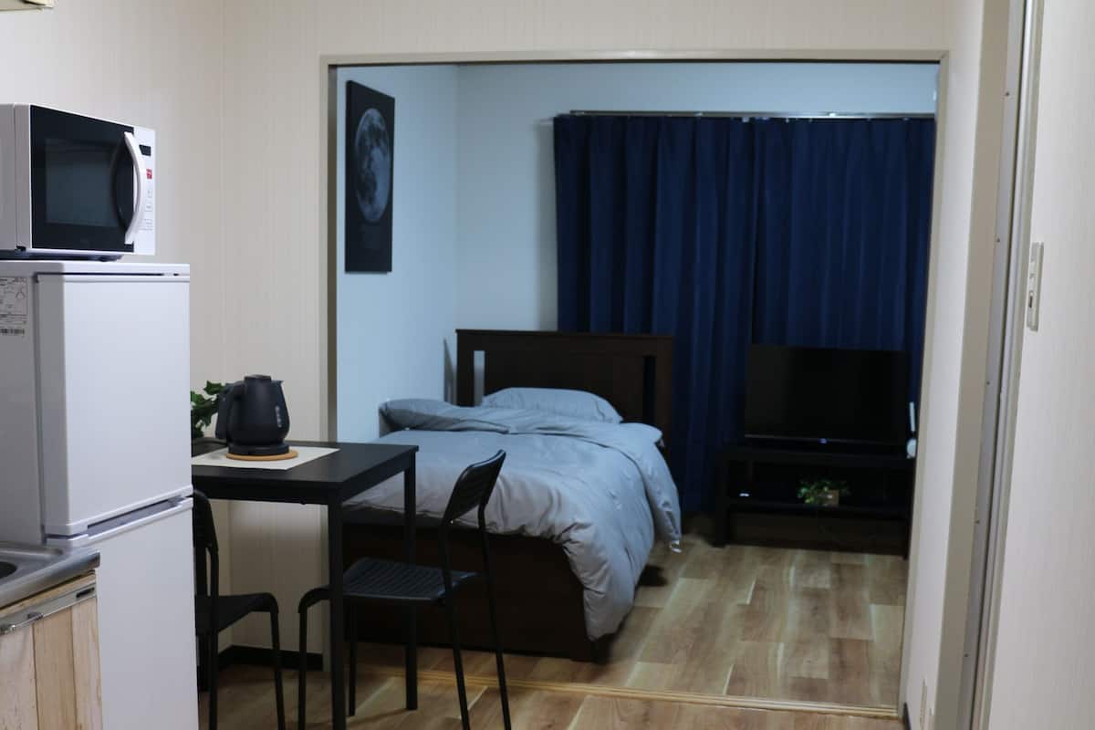 Une pièce compacte comprenant un coin cuisine, un lit avec un rideau bleu, une télévision et un petit tapis.