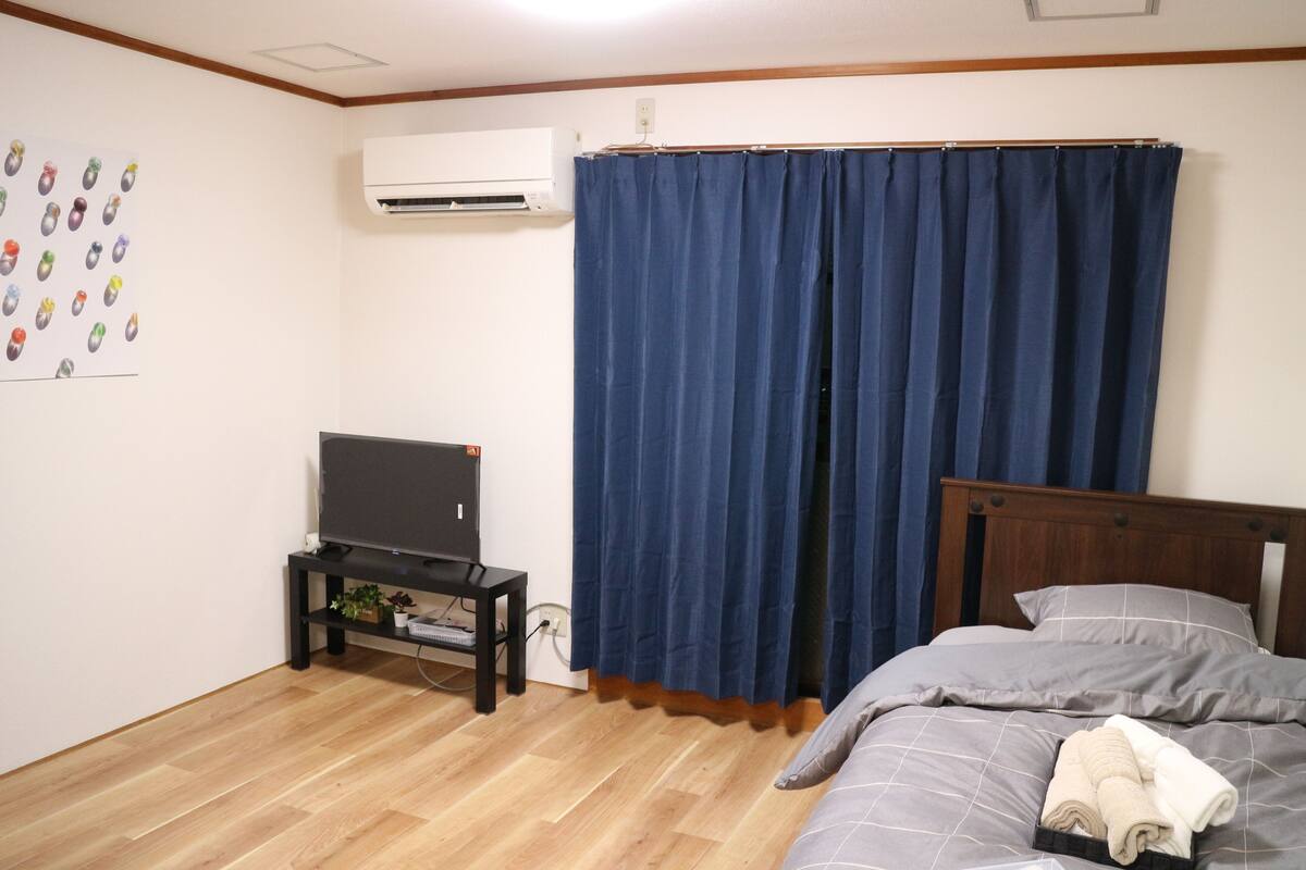 Un dormitorio con cama, televisor sobre un mueble, cortina azul y aire acondicionado instalado en la pared.