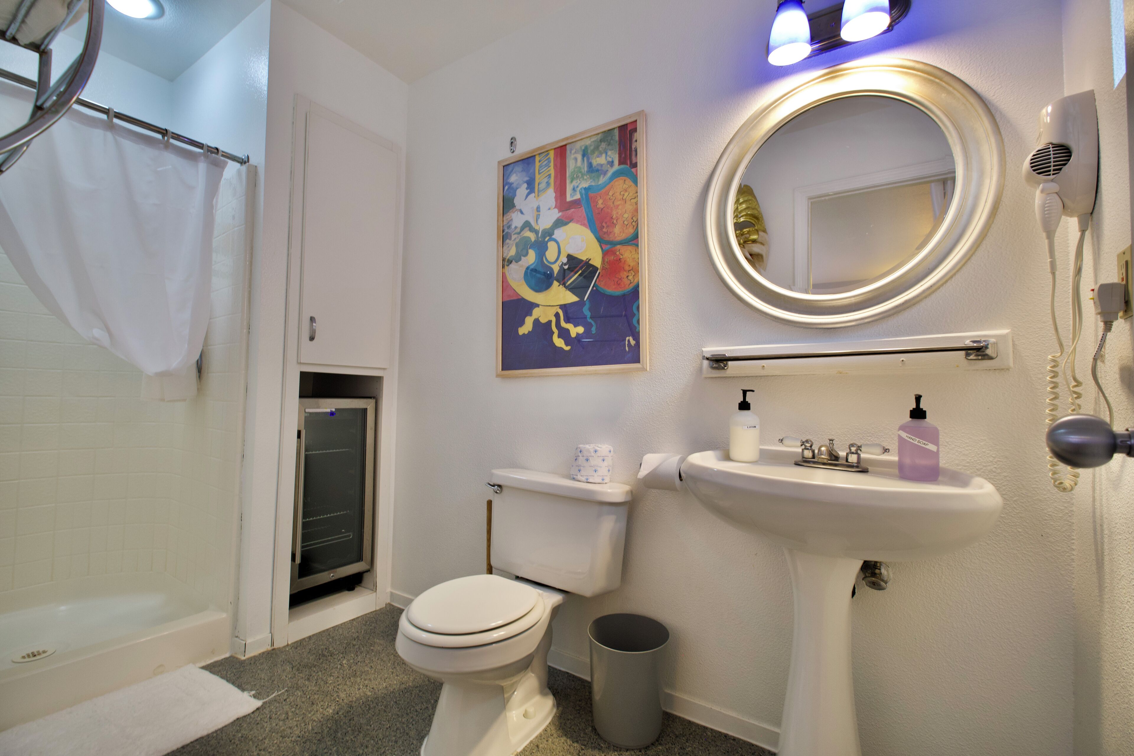 Une salle de bain avec des toilettes, un lavabo et une douche. Il y a un tableau accroché au mur et un sèche-cheveux également fixé au mur.