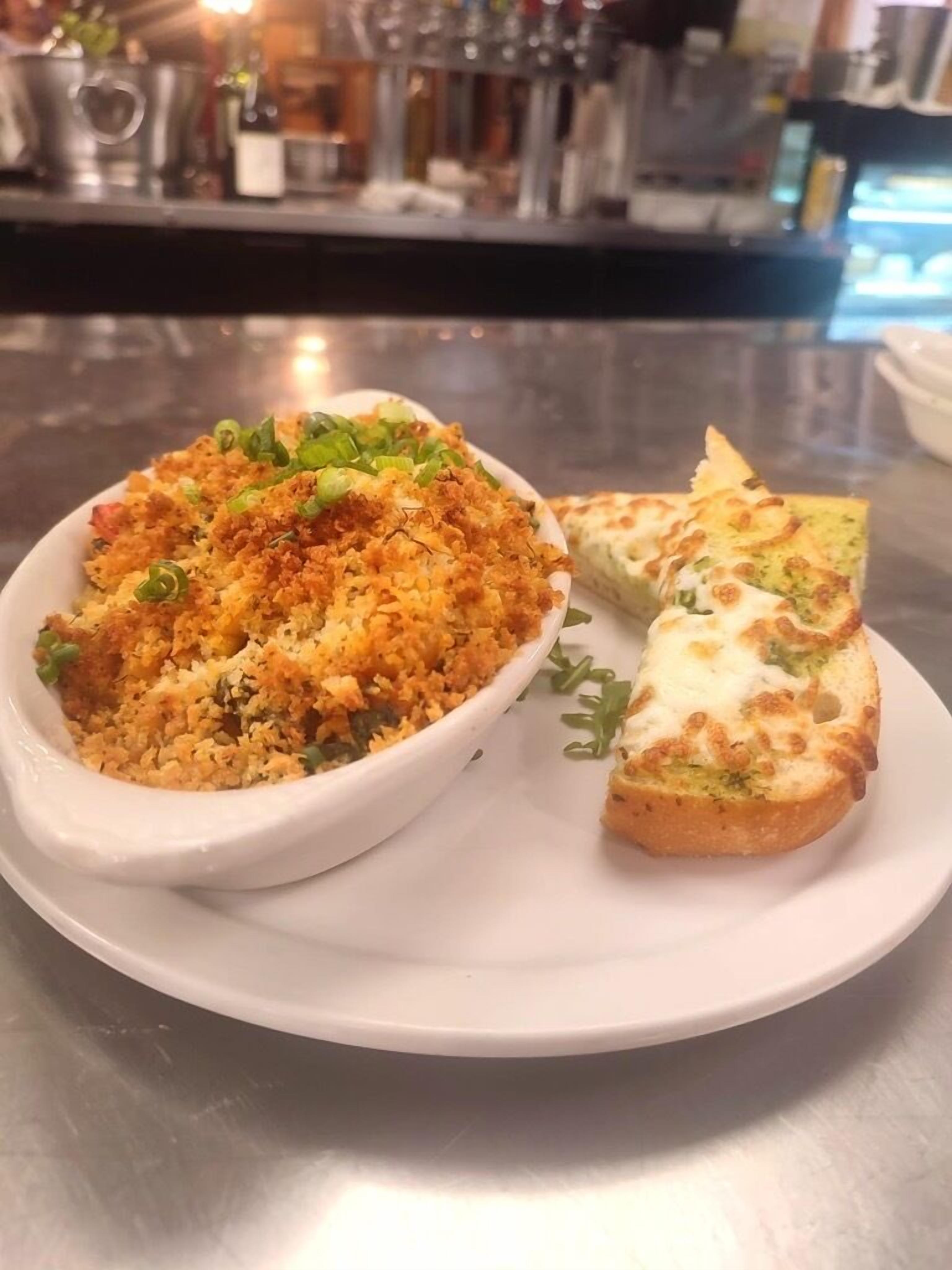 A white ceramic dish with a crumbly, golden-brown topping, possibly a casserole, accompanied by a slice of bread topped with melted cheese and green herbs.