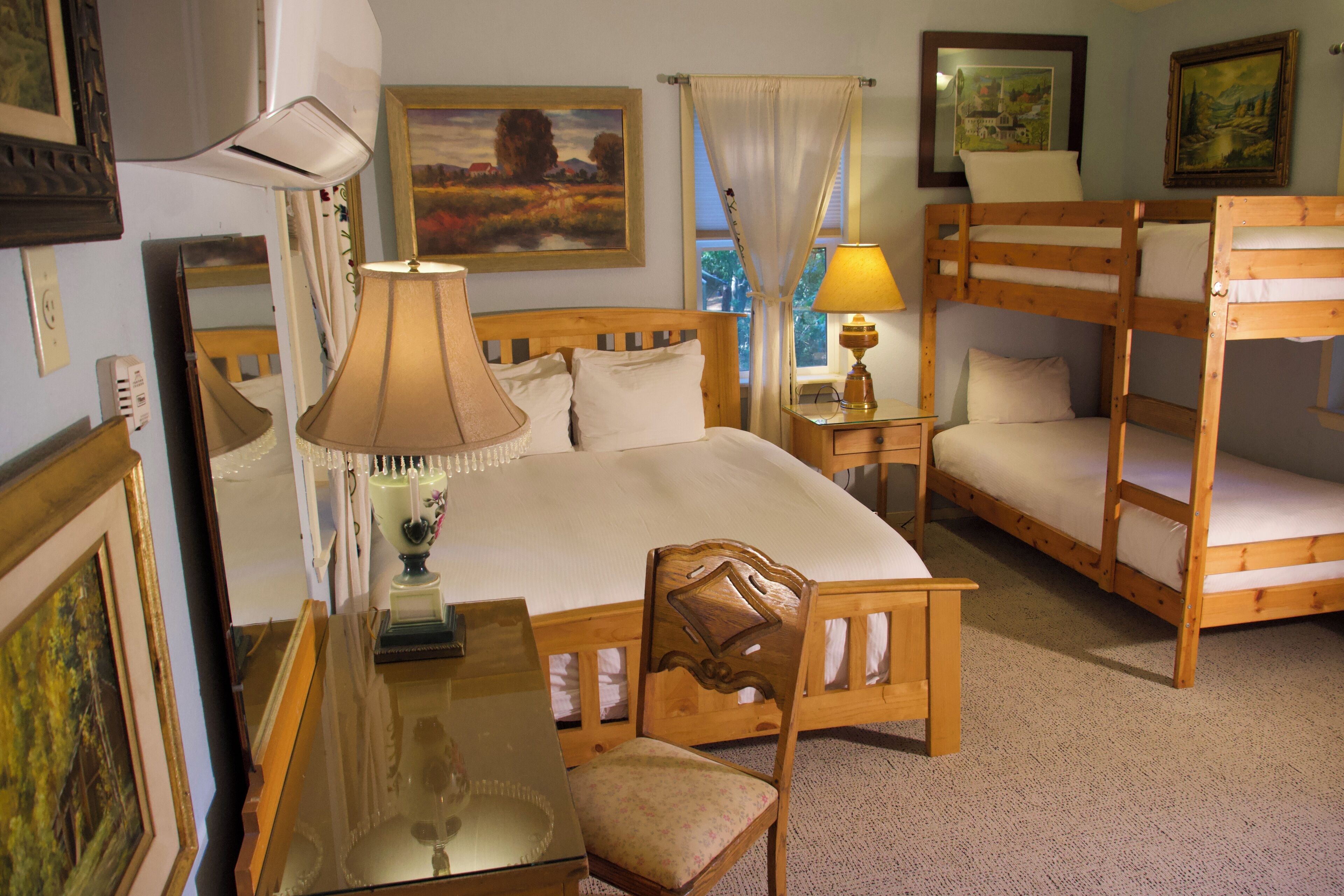 A bedroom with a wooden bed, a bedside table with a lamp, a chair, bunk beds, and framed pictures on the walls.