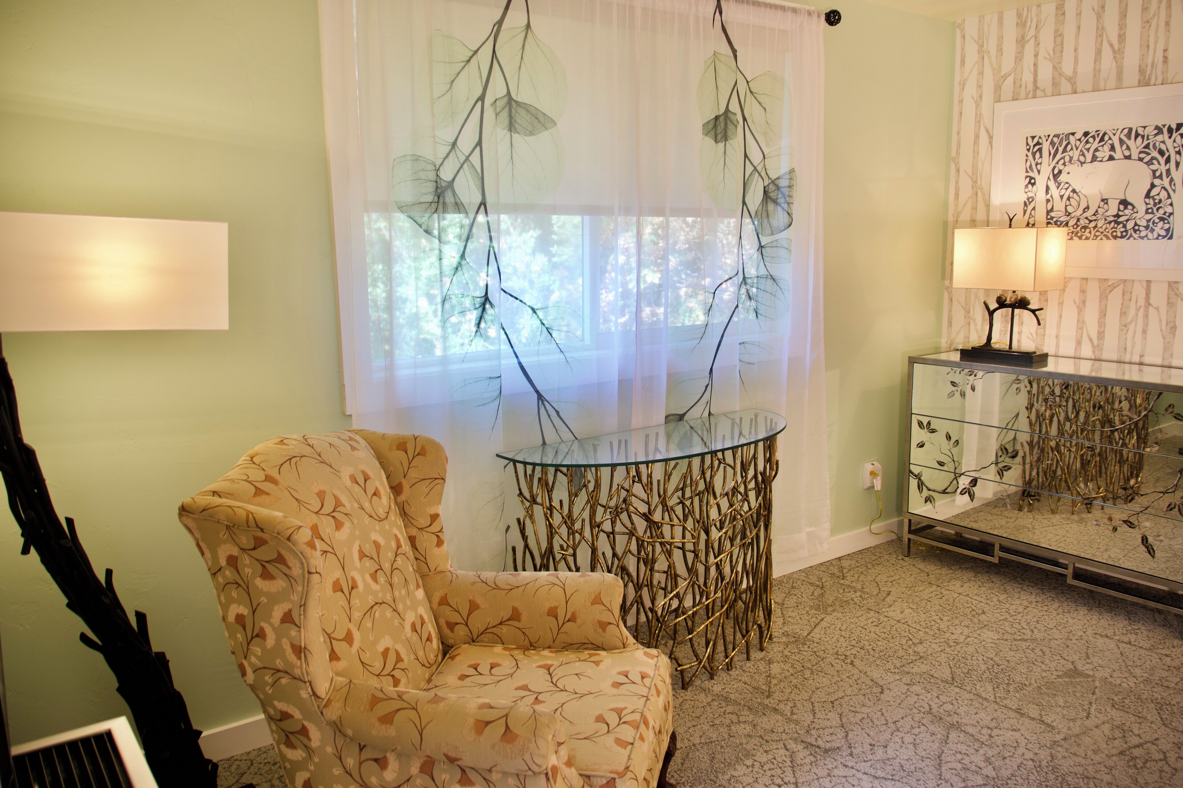 A room with a patterned armchair, a glass table with a decorative base, a lamp, and a framed artwork on the wall.
