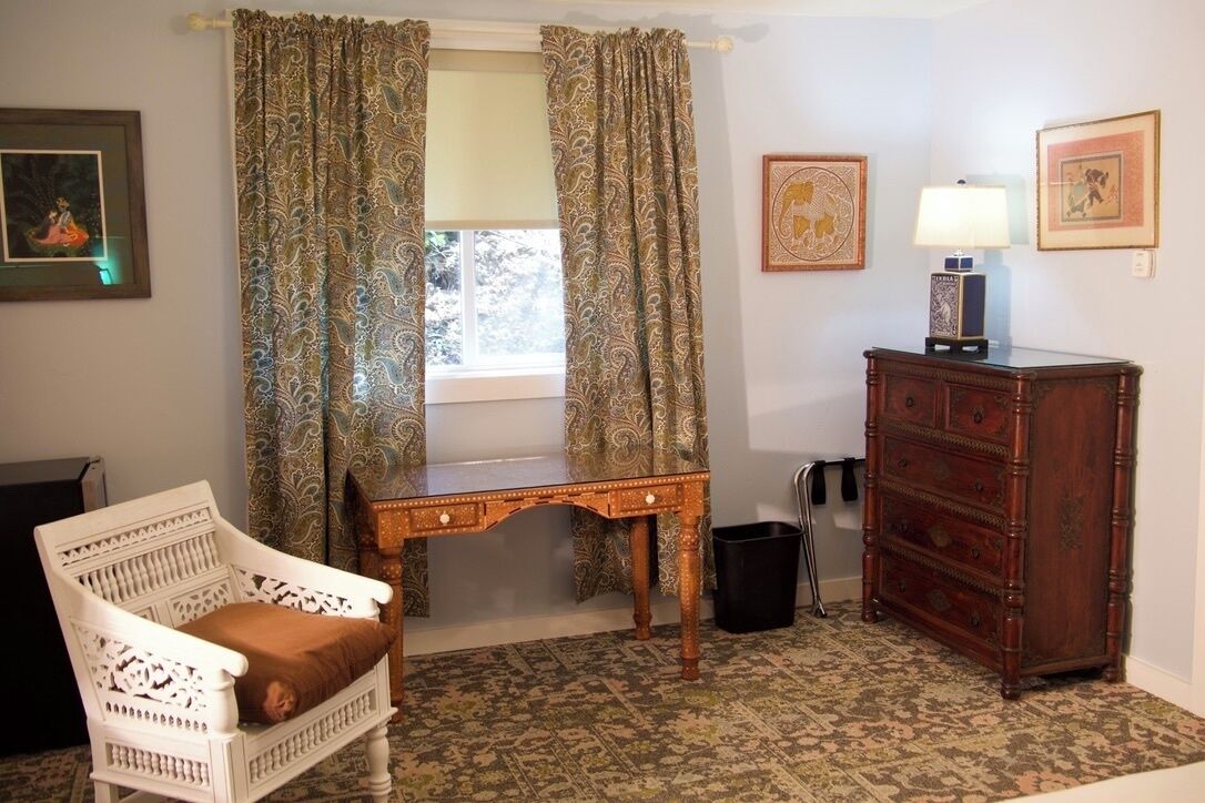 A room with a white chair, a wooden desk, a dresser, and patterned curtains.