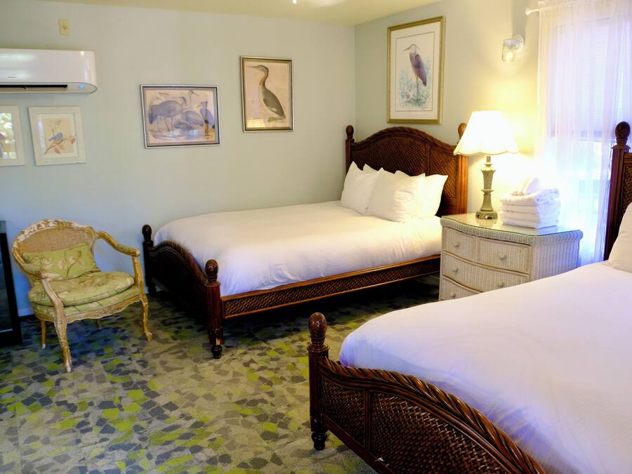 A hotel room with two single beds, a wooden headboard, a bedside table with a lamp, a chair, and a wall-mounted air conditioner.
