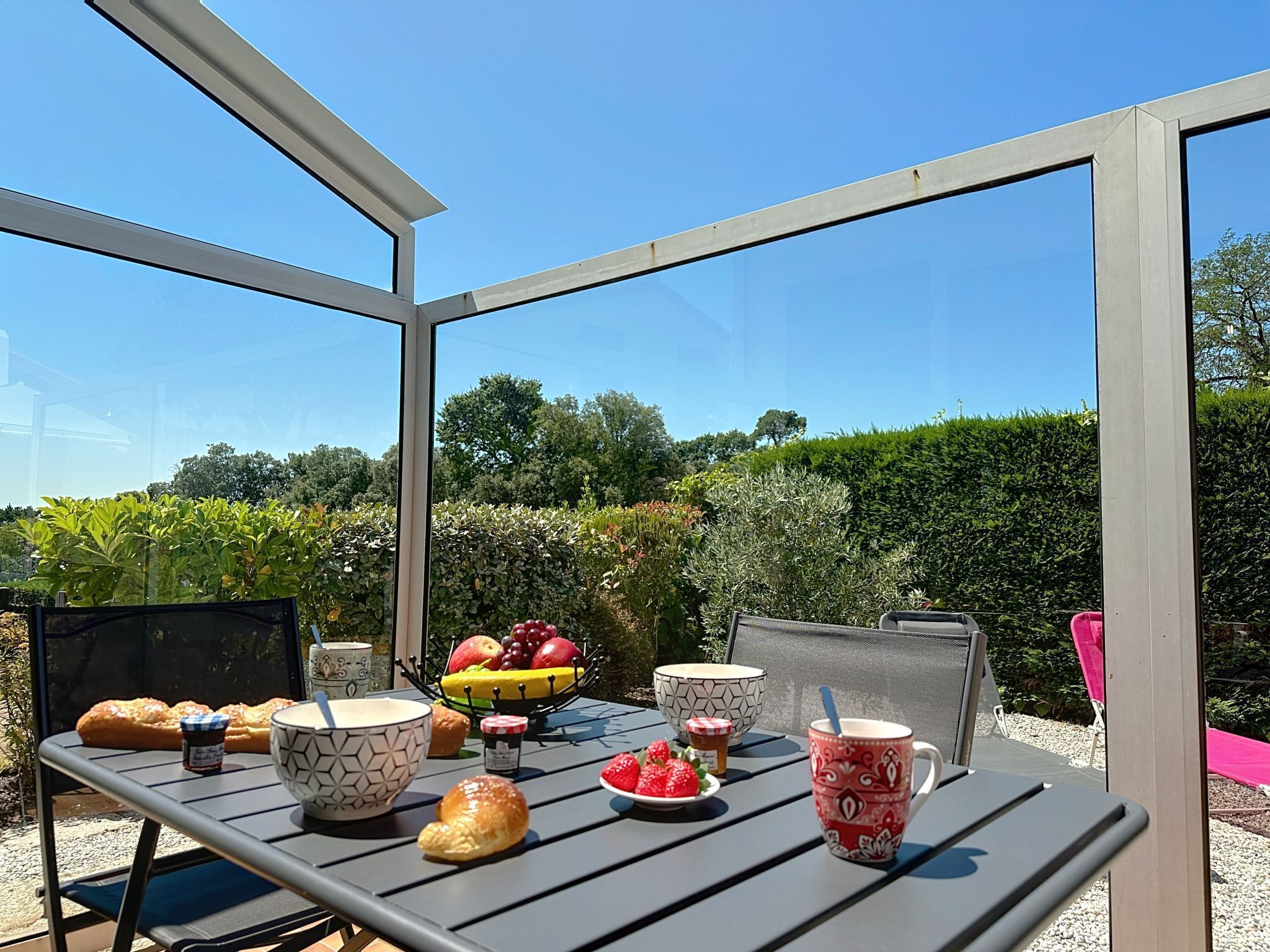 Mesa posta ao ar livre, com proteção de vidro, diversos alimentos e vista para o jardim.