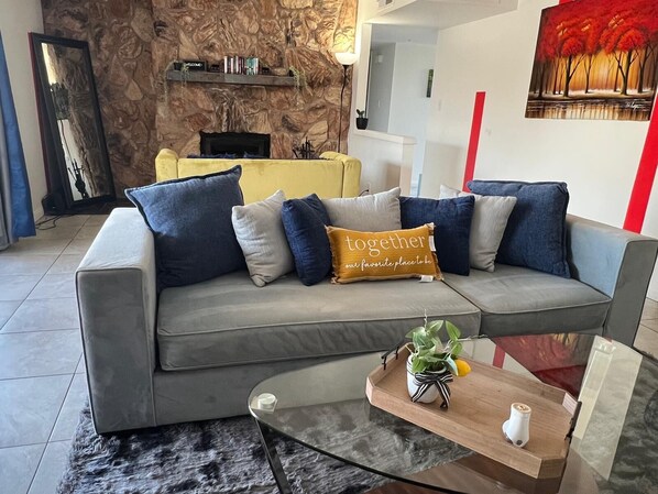 Un salon moderne avec un canapé gris, des coussins bleus et beiges, une table basse en verre et un tableau accroché au mur.