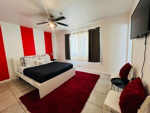 Une chambre à coucher avec un lit, un tapis rouge, une chaise blanche, un ventilateur de plafond et un téléviseur fixé au mur.