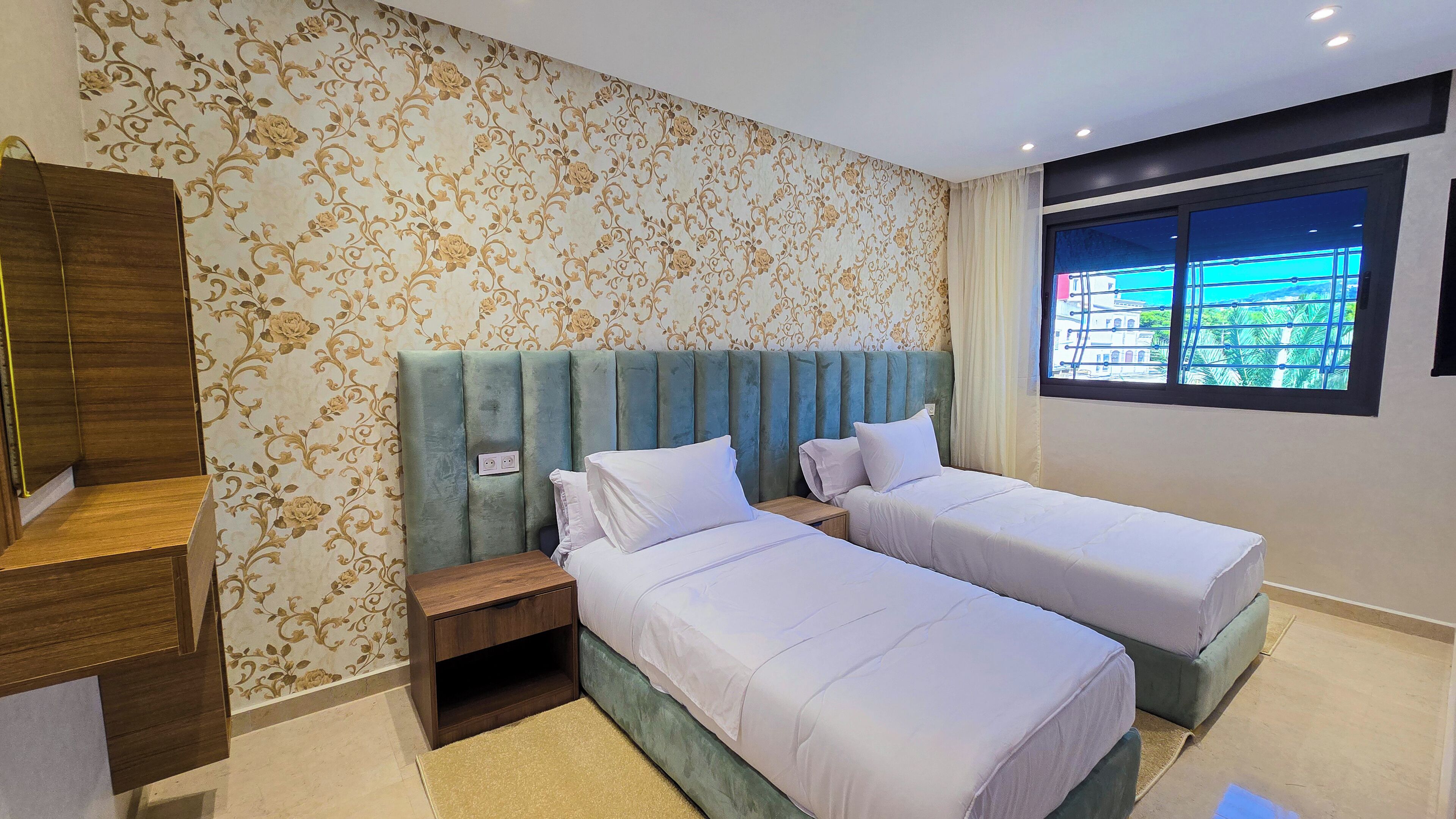 Hotellihuone, jossa on kaksi erillistä sänkyä, puinen vaatekaappi, pieni yöpöytä ja ikkuna, josta avautuu näkymä palmuille.