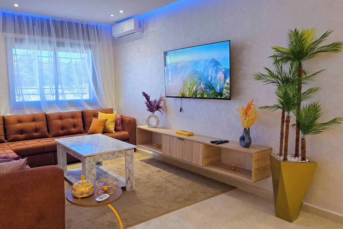 A modern living room with a brown sofa, a coffee table, a TV mounted on the wall, and a large window with sheer curtains.
