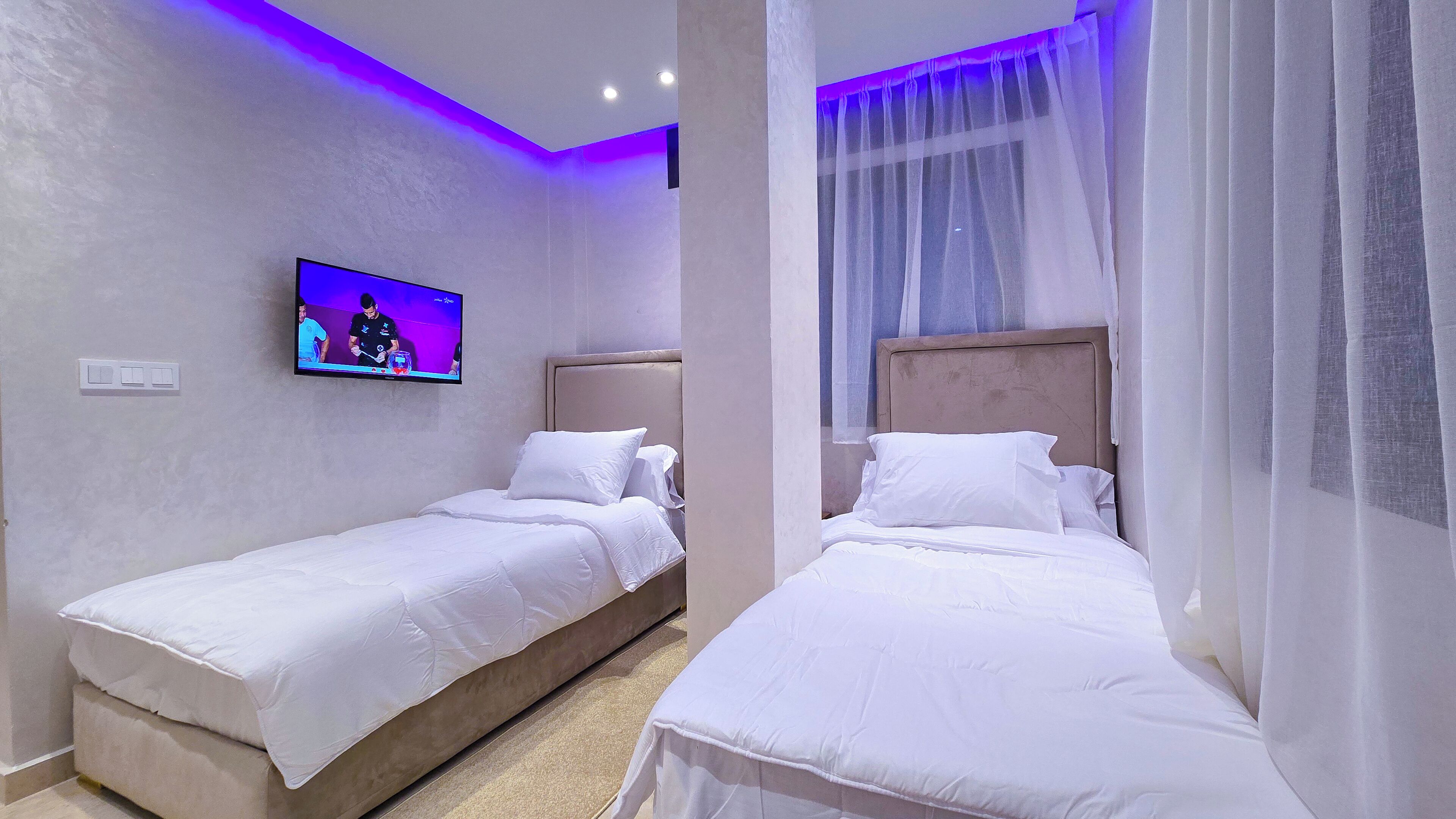 Een hotelkamer met twee enkele bedden, een aan de muur bevestigde televisie en een raam met dunne gordijnen.