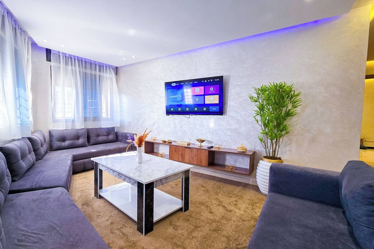 A modern living room with a sectional sofa, a marble coffee table, a flat-screen TV mounted on the wall, and a potted plant.