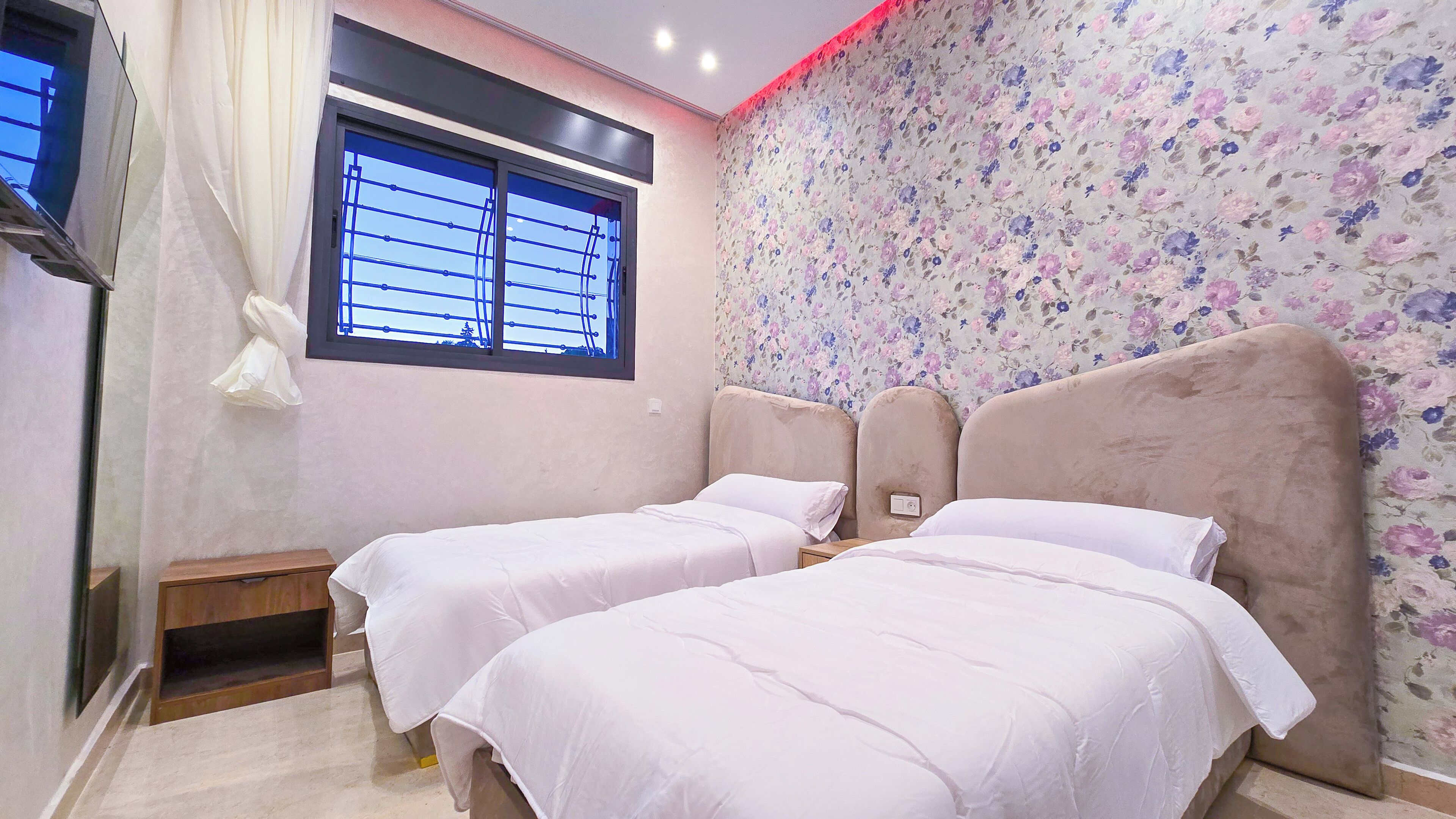 A bedroom with two beds, a window with blinds, a floral wallpaper, and a red light strip on the ceiling.
