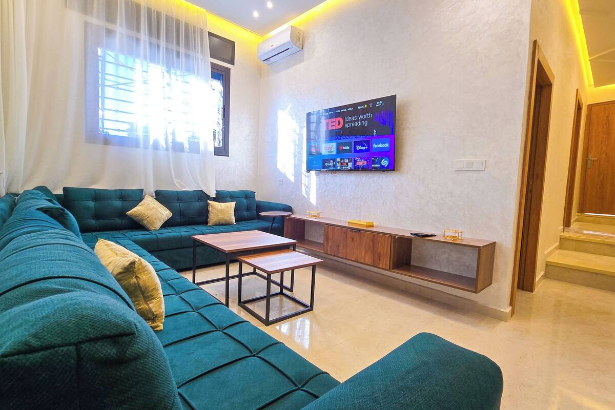 A modern living room with a teal sofa, a coffee table, and a TV mounted on the wall displaying various channels.
