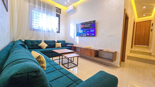 Una sala de estar moderna con un sofá color turquesa, una mesa de centro y un televisor de pantalla plana montado en la pared mostrando varios logotipos de redes sociales.