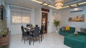 A modern living room with a green sofa, a dining table with chairs, and a kitchen area with a wooden cabinet and potted plants.