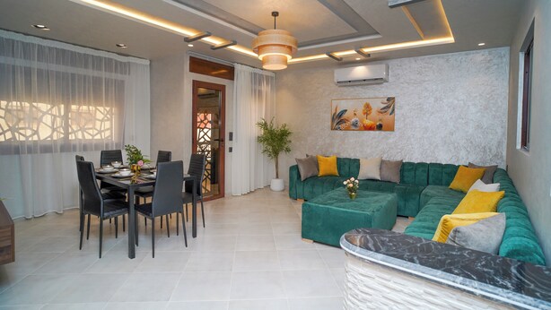 A modern living room with a dining area, featuring a green sofa, a dining table with chairs, and a ceiling with recessed lighting.