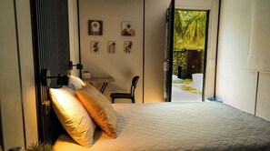 Un dormitorio con una cama, un escritorio y una silla. Hay obras de arte en las paredes y una puerta que da a un espacio exterior.