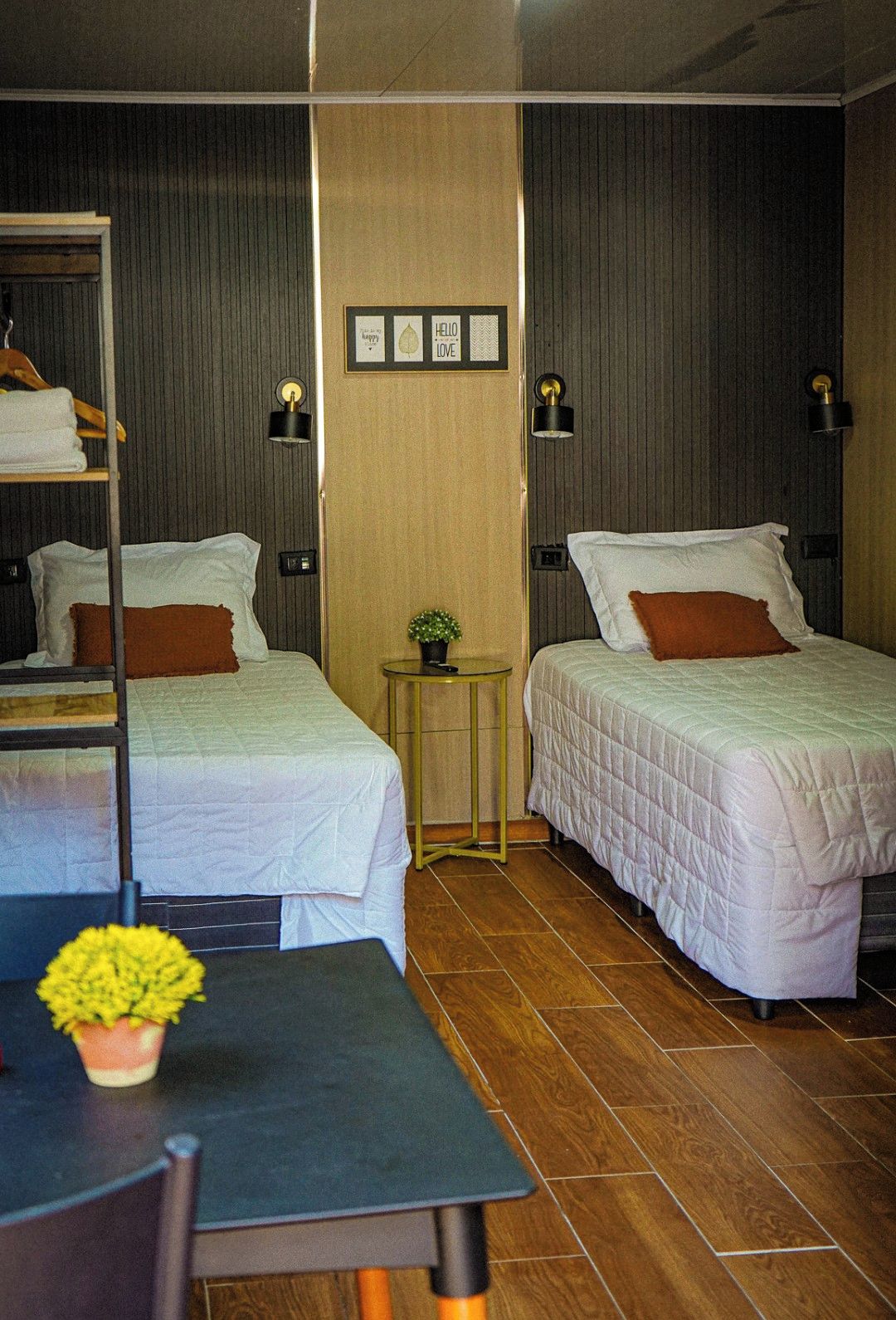 Une chambre d’hôtel avec deux lits, une petite table et une chaise. On y trouve une étagère avec des serviettes et un pot de fleurs sur la table.