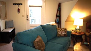Un salon chaleureux avec un canapé de couleur turquoise, une table d’appoint en bois sur laquelle se trouve une lampe, et une porte donnant sur un balcon.