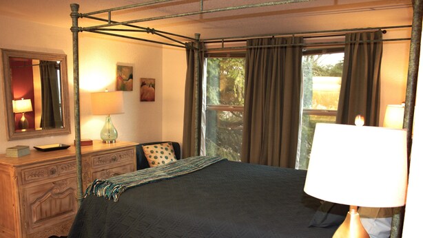 Un lit à baldaquin avec une couverture sombre, une commode en bois avec un miroir, une lampe et un tableau accroché au mur.