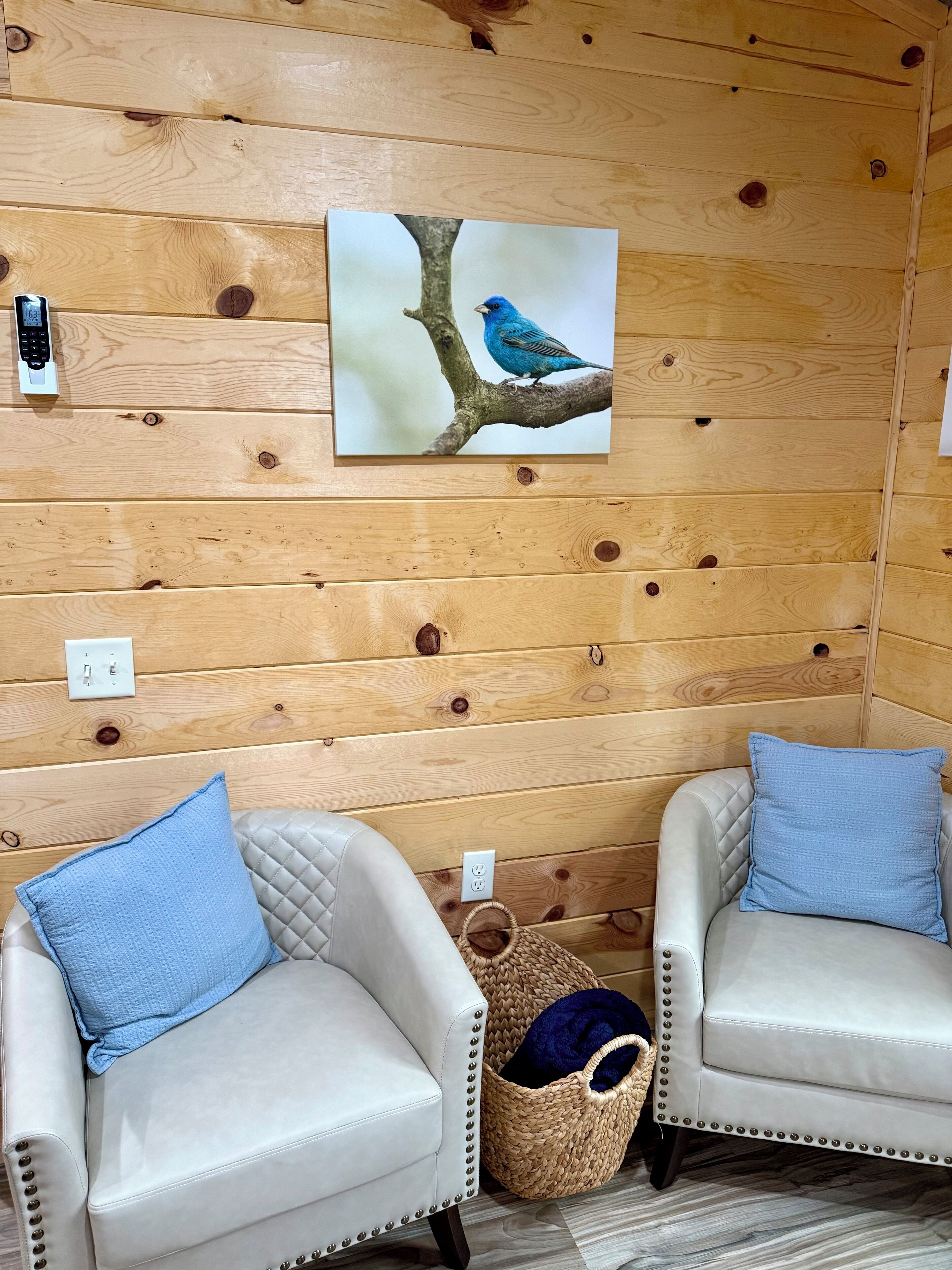 A room with wooden panel walls, two white armchairs with blue cushions, a wall-mounted painting of a bird, a wall socket, and a woven basket.
