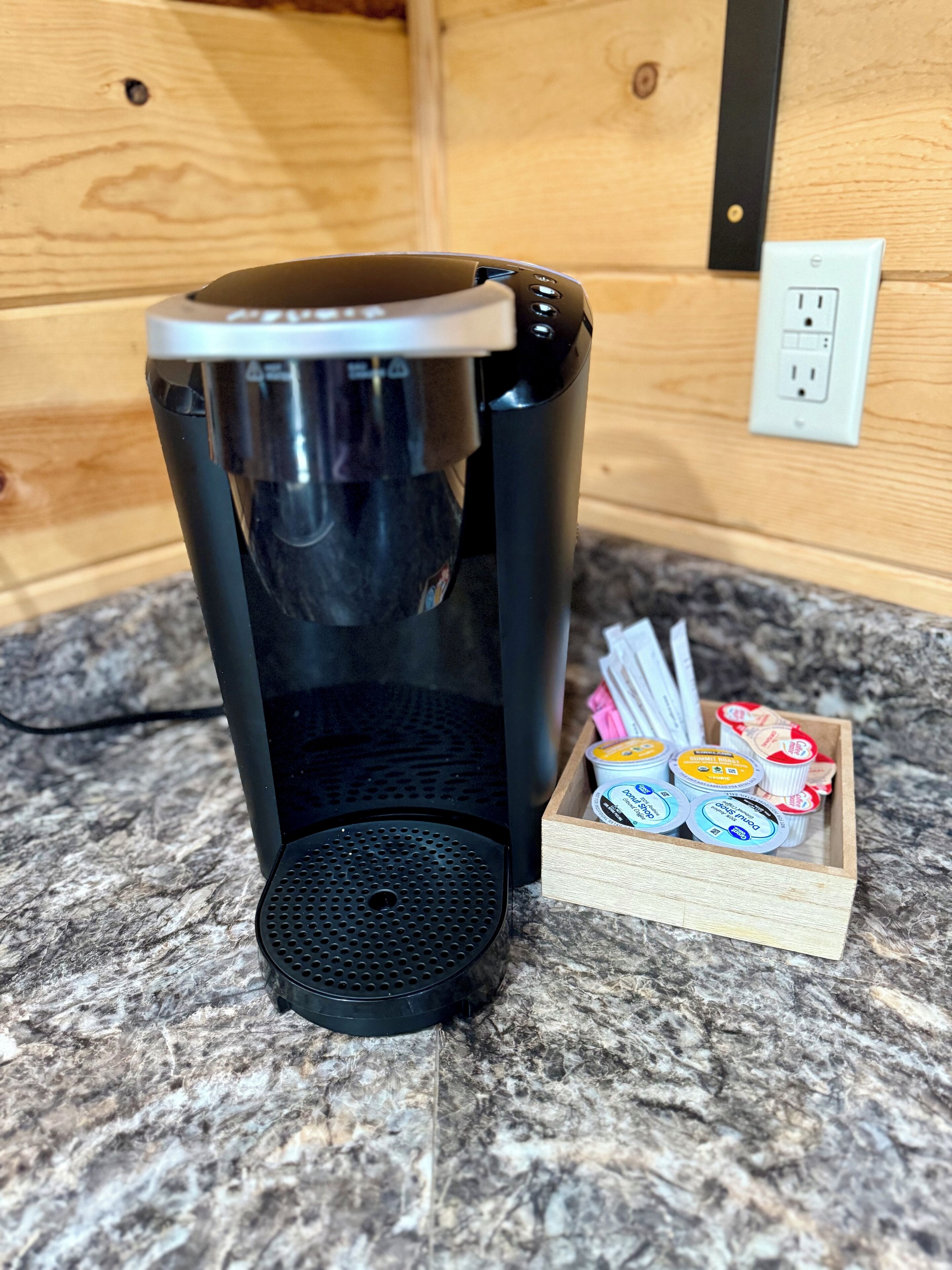 A coffee maker on a granite countertop next to a wooden box with yogurt containers.