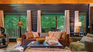 A cozy living room with a brown sofa, green armchair, and a coffee table with a bowl and books. Large windows with curtains provide a view of trees outside.