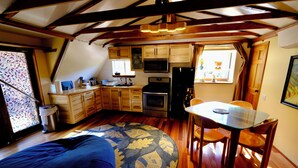 A compact kitchen with wooden cabinets, a microwave, and a refrigerator. There's a dining table with chairs and a window with curtains.