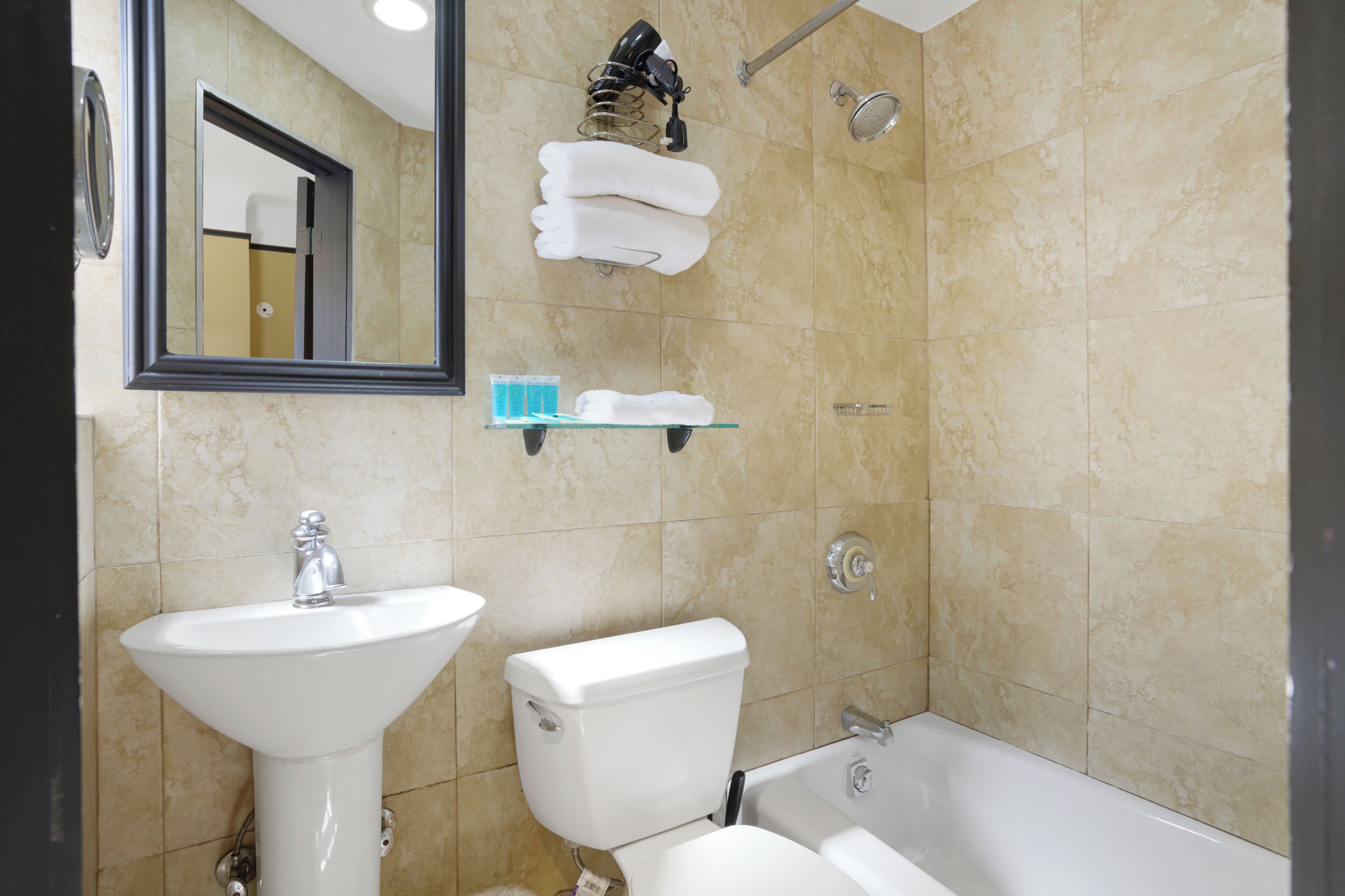 Ein Badezimmer mit Waschbecken, Toilette und Badewanne. Handtücher und Toilettenartikel liegen auf einem Regal.