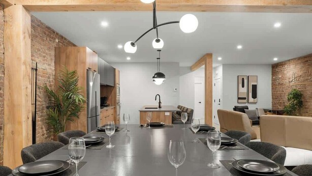 Un comedor moderno con una larga mesa dispuesta para seis personas, una cocina al fondo y una pared de ladrillo.