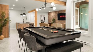 Una mesa de billar con sillas, una zona de cocina, una mesa de comedor con sillas, un televisor mostrando 'NETFLIX' y un ventanal grande.