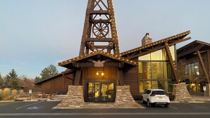 Un bâtiment de style rustique, doté d’une tour imposante, de grandes fenêtres et d’un espace de stationnement où se trouve une voiture blanche.