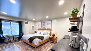 Une chambre d’hôtel moderne équipée d’un lit, de tables de chevet, d’un bureau, d’une chaise, d’une plante en pot et d’une fenêtre donnant sur un paysage agréable.