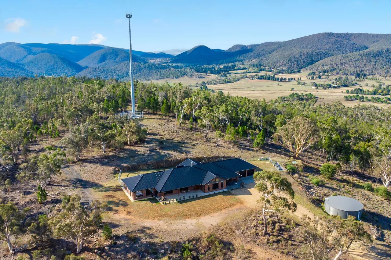 A large, single-story building with a dark roof, surrounded by a natural landscape with trees and hills in the background.