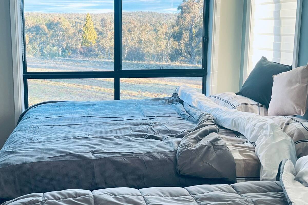 A bedroom with a large window, a bed with a grey bedspread, and pillows.