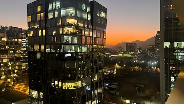 A modern high-rise building with multiple balconies and large windows, surrounded by other buildings and a mountain in the background.