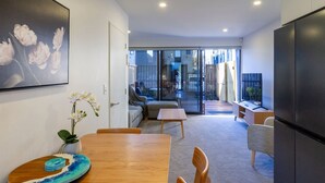 A modern kitchen with a wooden dining table, chairs, and a refrigerator. There is a glass door leading to a balcony with a view of the outside.