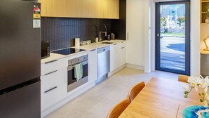 A modern kitchen with a built-in oven, a dishwasher, and a refrigerator. There is a dining area with a wooden table and chairs.