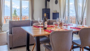 Un coin repas avec une table en bois, des chaises à revêtement gris et une bouteille de vin. Des rideaux sont accrochés aux fenêtres, et une horloge est visible au mur.