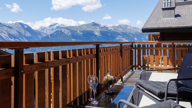 Un balcon avec une balustrade en bois, une table sur laquelle se trouvent un globe terrestre et des verres à vin, et une vue sur des montagnes enneigées.