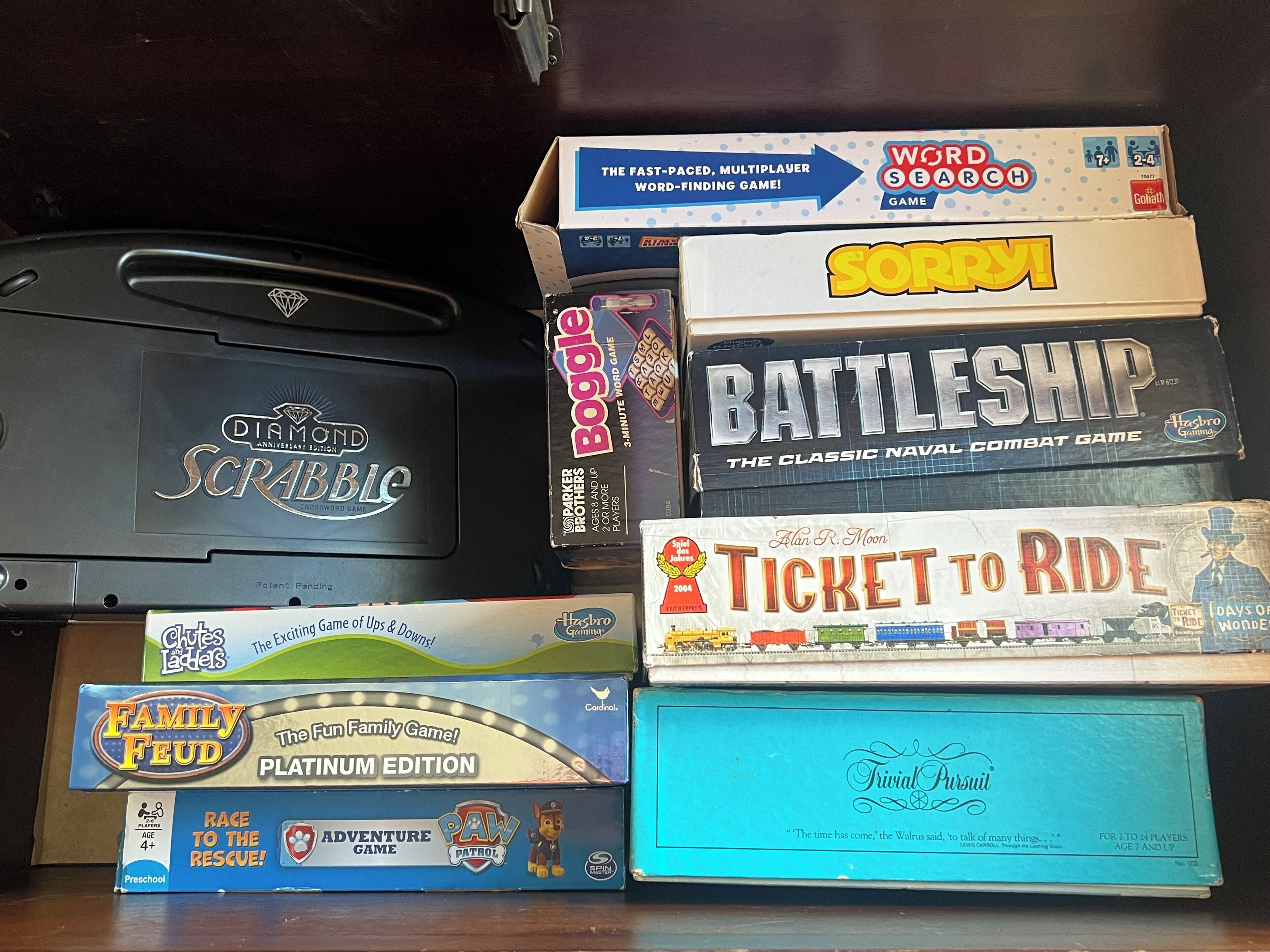 A collection of board games including Scrabble, Sorry!, Battleship, Ticket to Ride, and others, arranged on a shelf.