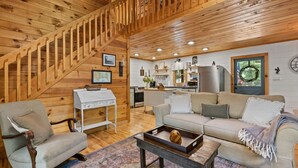 A cozy cabin interior with a wooden staircase, a living room with a sofa and armchair, a kitchen area, and a dining table with a decorative bowl. - Walk to Lake Blue Ridge | Hot Tub + Fire Pit (Morganton)