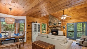 A cozy cabin interior with a stone fireplace, wooden walls, and a dining area with a table and chairs. - Walk to Lake Blue Ridge | Hot Tub + Fire Pit (Morganton)