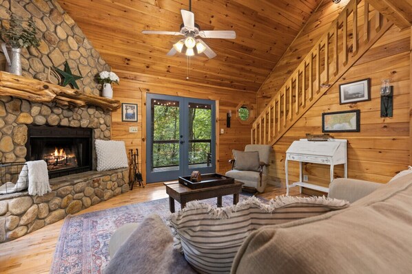 L’intérieur chaleureux d’un chalet avec une cheminée en pierre, un ventilateur de plafond, un escalier en bois et une grande fenêtre donnant sur un paysage verdoyant.