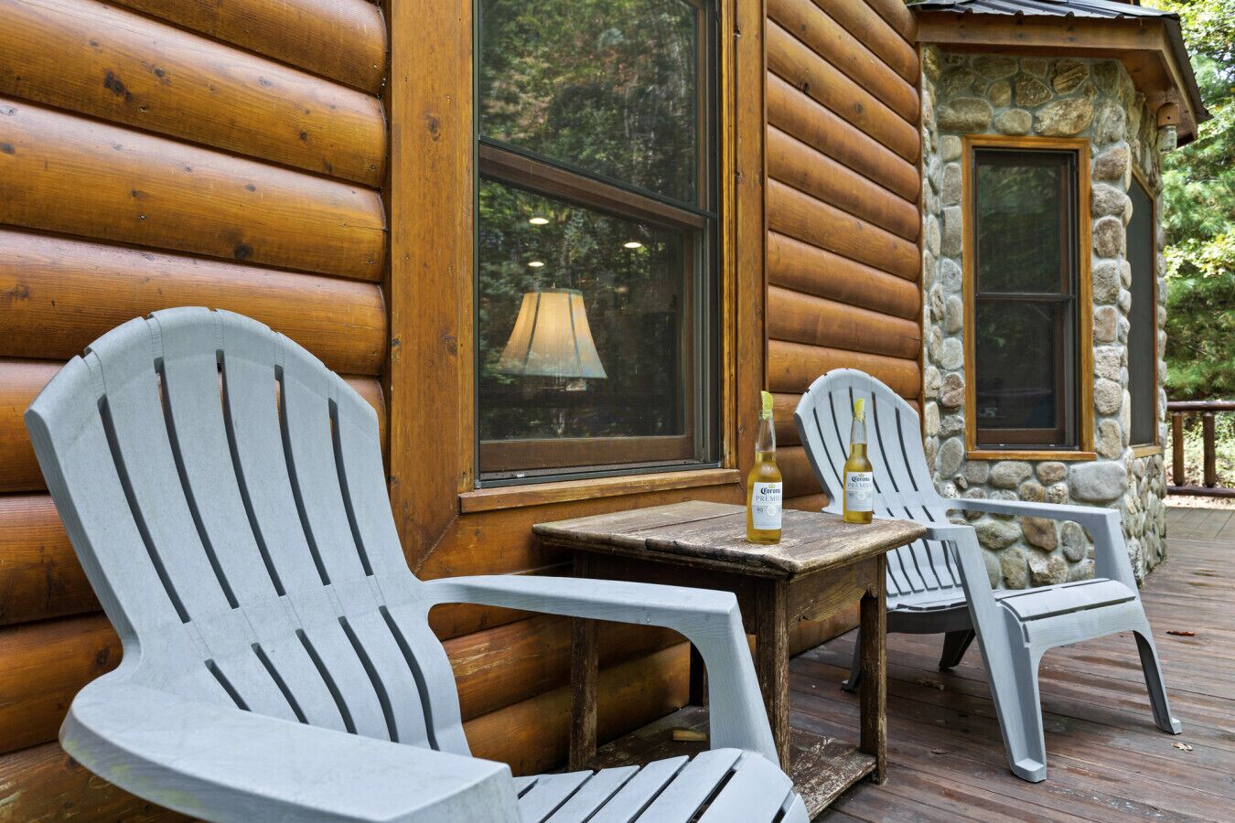 A wooden cabin with two Adirondack chairs and a table, a bottle of Corona beer, and a stone wall.