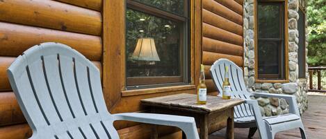 A wooden cabin with two Adirondack chairs and a table, a bottle of Corona beer, and a stone wall.